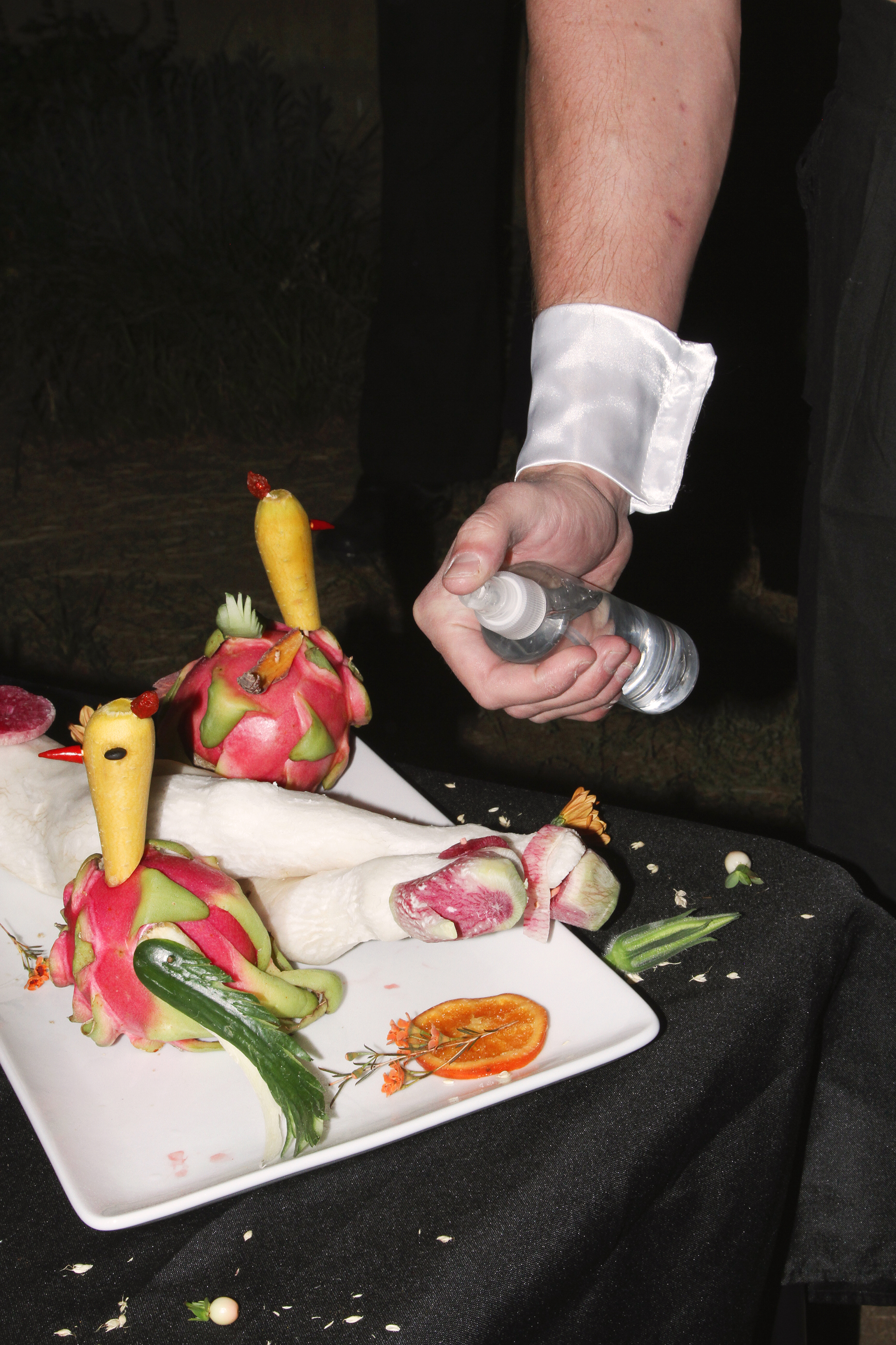 Detail of a shirtless white male butler spraying water on fruit sculptures on a black table
